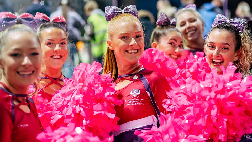 Cheerleaders are stood smiling holding their Poms, in Loughborough colours, Pink and Purple.