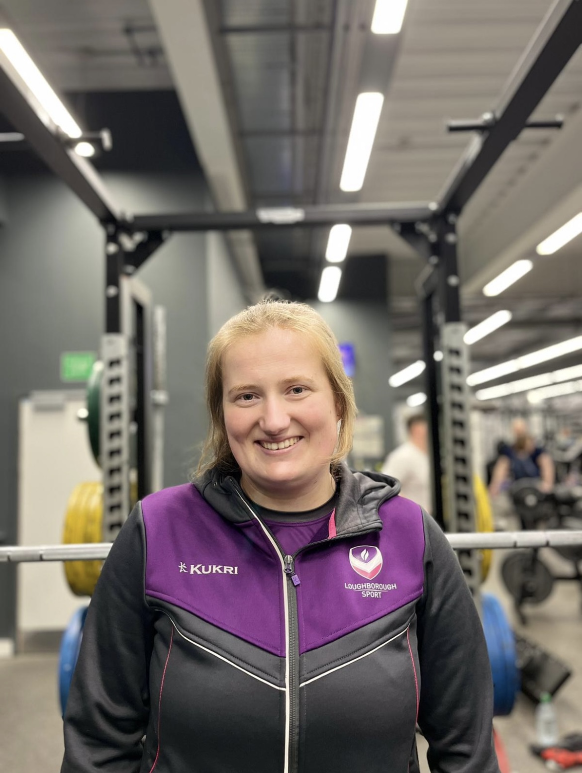 Fitness Coach, Joanna, smiling in Holywell Fitness Centre with gym equipment in the foreground