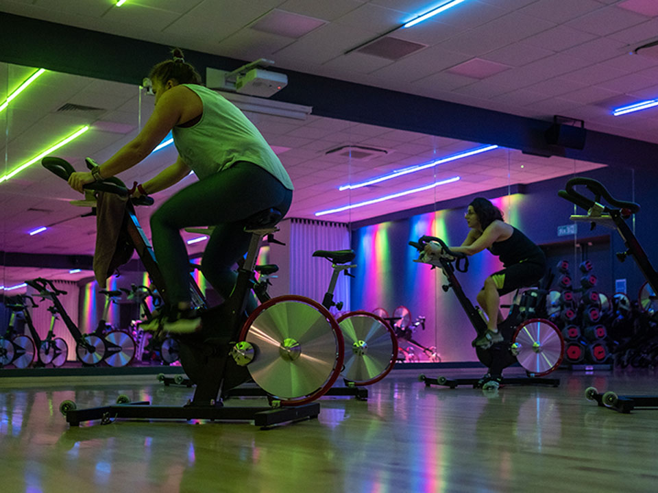 Two people ride stationary bikes in a fitness studio with colourful neon lights reflected on the walls and floor.