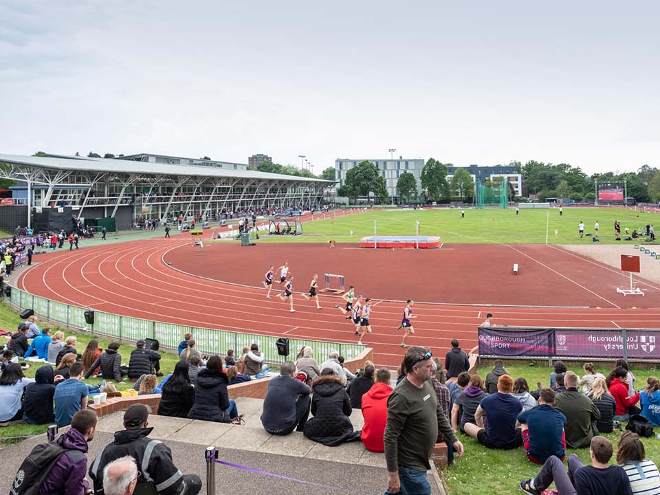 In the sunshine, there are athletes running on the track at a meet, with spectators watching on.