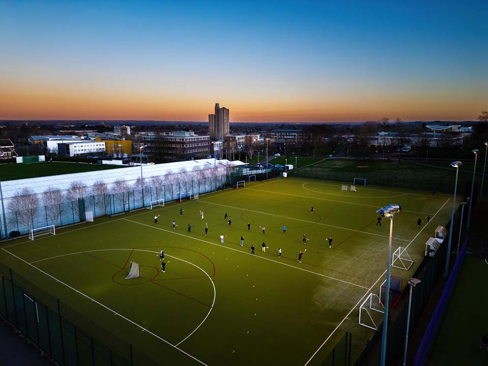 A sand based pitch under floodlights has people playing sport on it.