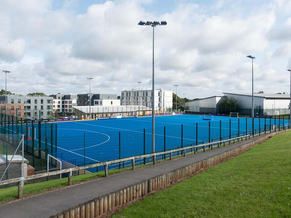 A blue water based hockey pitch sits behind fencing.