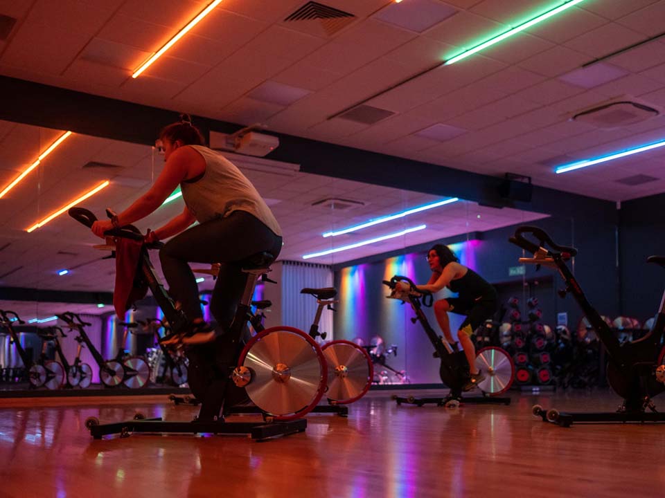 A group of members are taking part in a spin class, in a studio with a wall of mirrors and lights.