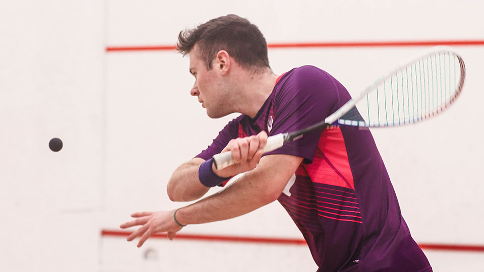 A male squash player wearing Loughborough kit and about to hit the ball