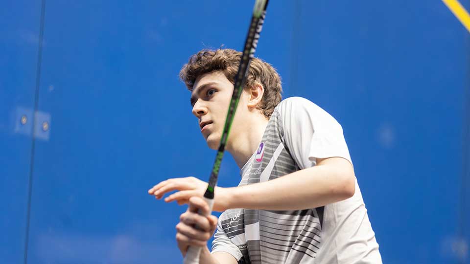 A male squash player wearing Loughborough kit and holding a racket about to hit the ball during a training session