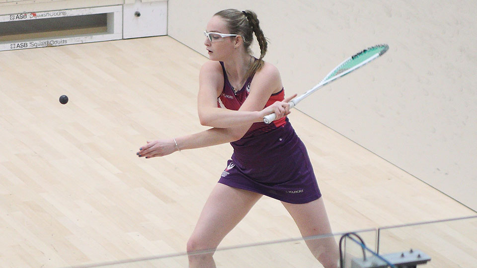 A female squash player wearing Loughborough kit and holding a racket about to hit the ball during a training session