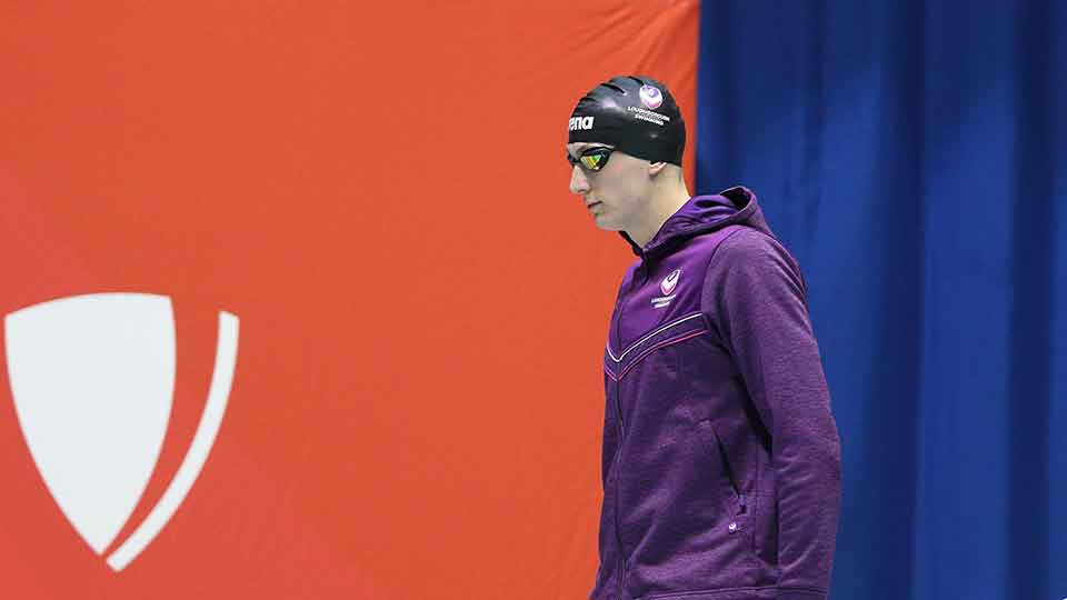 Luke, wearing a swimming cap, goggles and purple Loughborough Swimming jacket whilst standing side on to the camera.