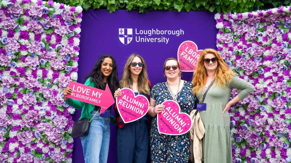 Four people standing in front of a flower wall at an alumni reunion event, holding banners reading 'Alumni Reunion' and 'Loughborough Family'.
