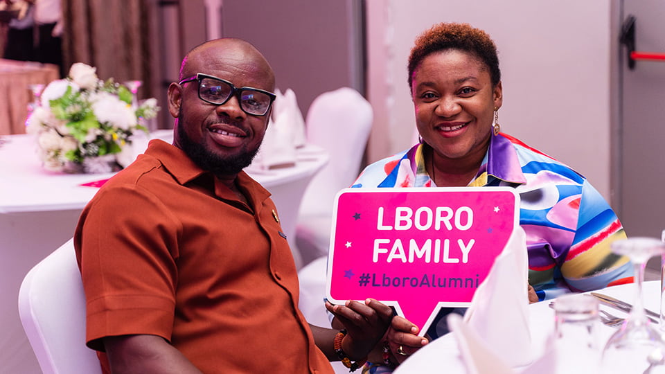 Two people sat together at a table, one is holding a sign reading 'Loughborough Family #LboroAlumni'.