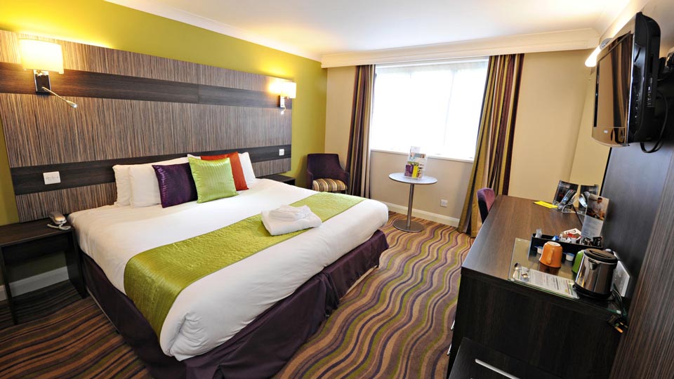 View of a double room: A modern dark brown wavy carpet, backed with a lime green wall and dark wood panelling behind the bed, which is covered in white linen, a lime green bed topper with multicoloured cushions; desk and chair to the right.