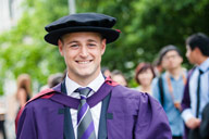 A smiling PhD graduand, wearing the trademark tasselled floppy cloth bonnet and purple gown with maroon edging and maroon hood.