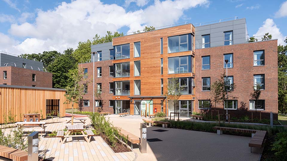 Claudia Parsons hall in brilliant sunshine - a four-storey accommodation block with visual interest from big windows, wood panelling, grey metallic cladding, and with wooden seating and landscape planting outside. 