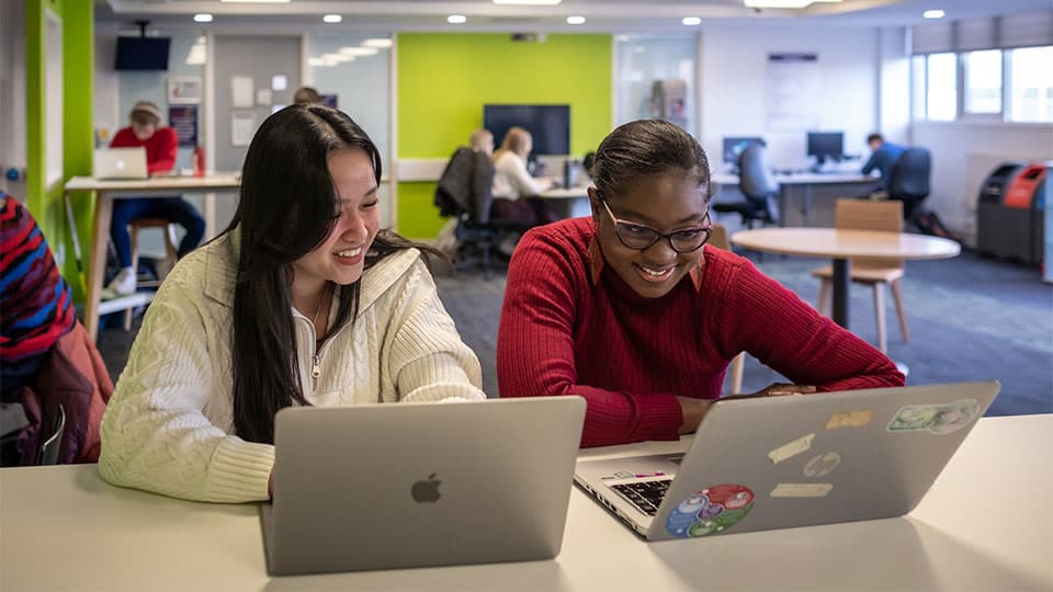 two students smiling and looking at computer screens in the library