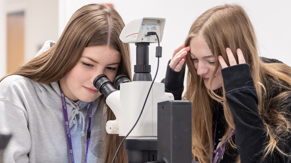 Two students sitting at a bench, one looking into a microscope