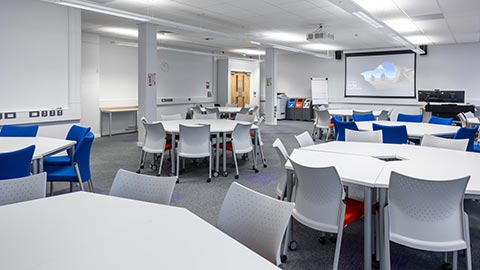 Large, modern classroom or training space with movable white tables arranged in groups, white and blue chairs on wheels, a projector screen at the front, and bright overhead lighting.