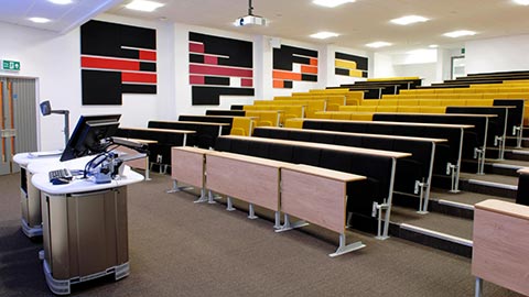 Large tiered lecture theatre with rows of yellow and black seats facing a lecturer’s podium equipped with computers and presentation controls.