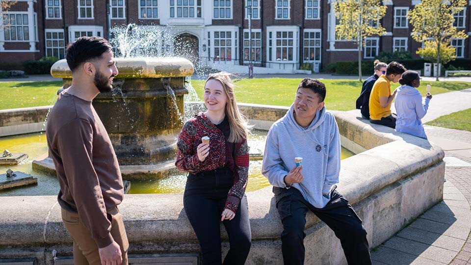 3 students in front of the Hazelrigg building sitting on the edge of the water fountain, laughing and having a conversation while holding ice creams.
