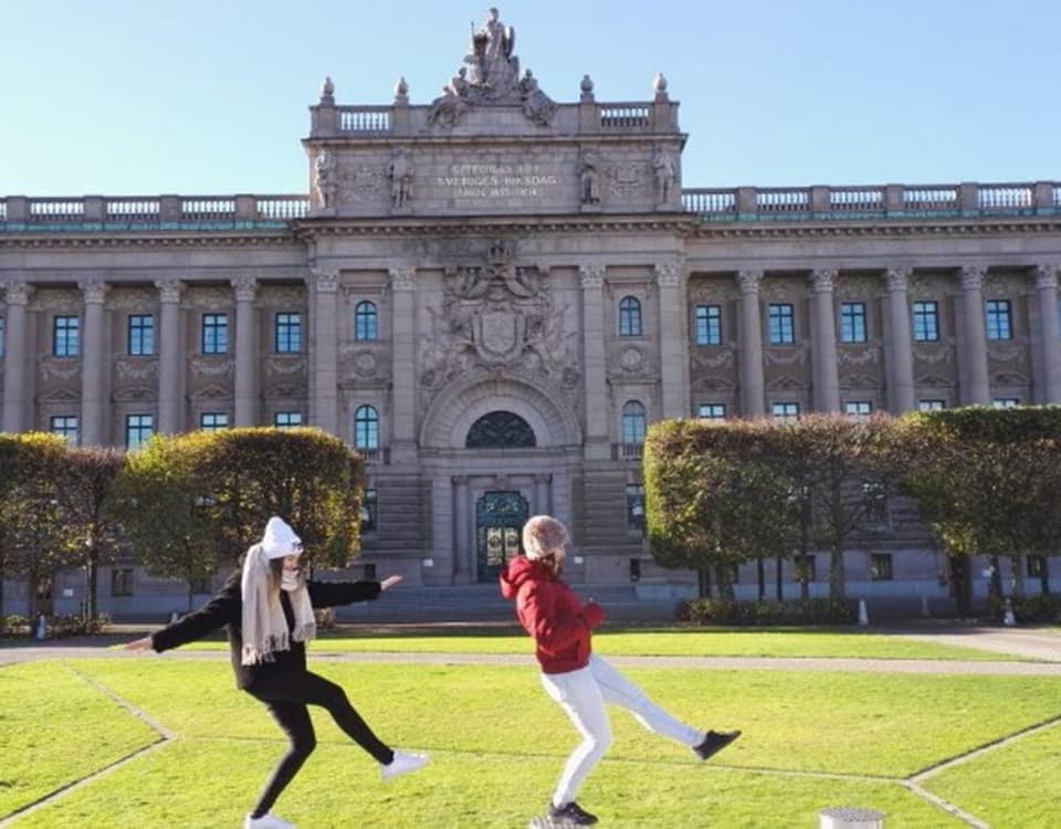 Two students on a grassy area outside the front of a large grand building. They are balancing on posts with one foot in the air.
