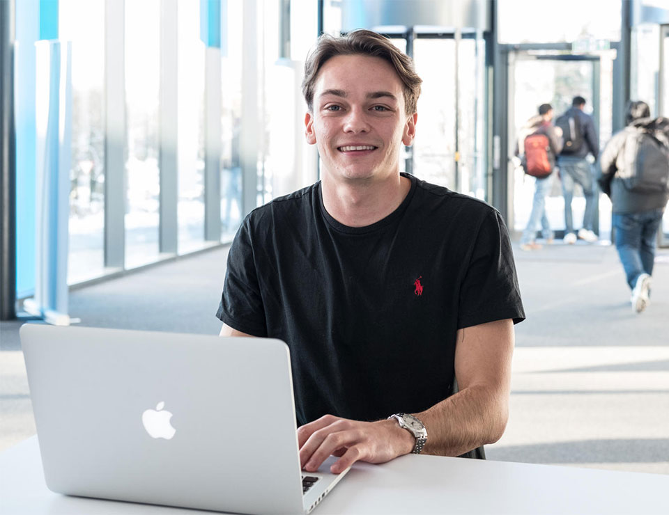 Male student sat at a desk with his hands on the keypad of a Macbook pro; he is looking directly at the camera whilst smiling.