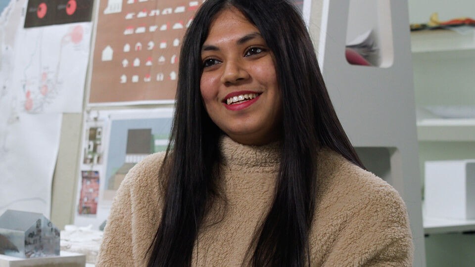 Female student sitting down on a chair in a built environment studio, smiling at someone.