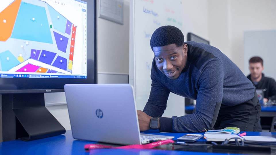 Student standing in the lecture room, leaning over a laptop, showcasing a construction project on a large screen on the wall