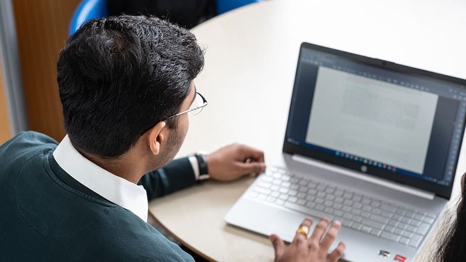 A male student sitting at a table, looking at his open laptop