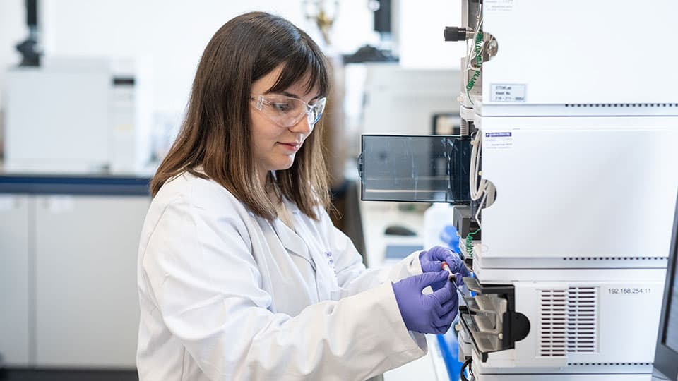 A female student wearing a white lab coat and goggles while performing a chemistry experiment