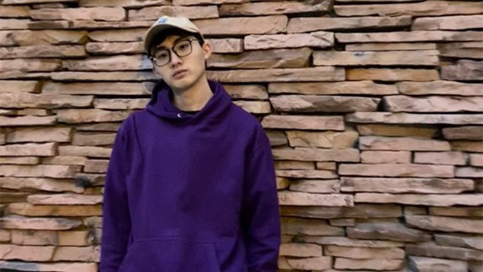 Jonathan, standing in front of a brick wall wearing a purple jumper and a baseball cap looking directly at the camera.