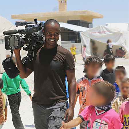 A dark-skinned man wearing a brown t-shirt and carrying a professional video camera on his shoulder, accompanied by a group of children