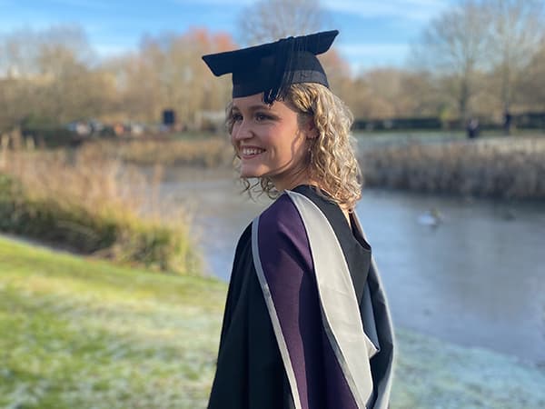 Olivia in a graduation gown by the lake at Loughborough University.