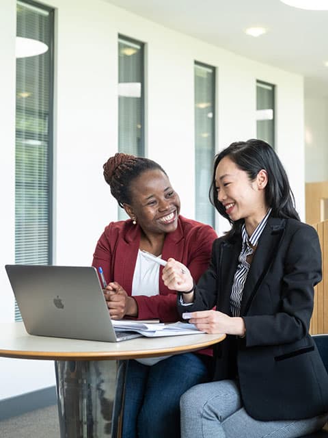 Two students sitting at a table with a laptop chatting and laughing.
