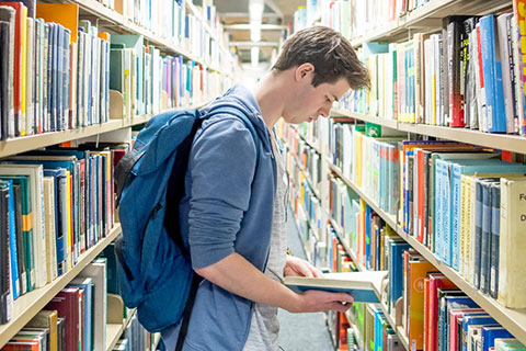 A male student wearing a rucksack standing between two bookshelves in the Library browsing a book