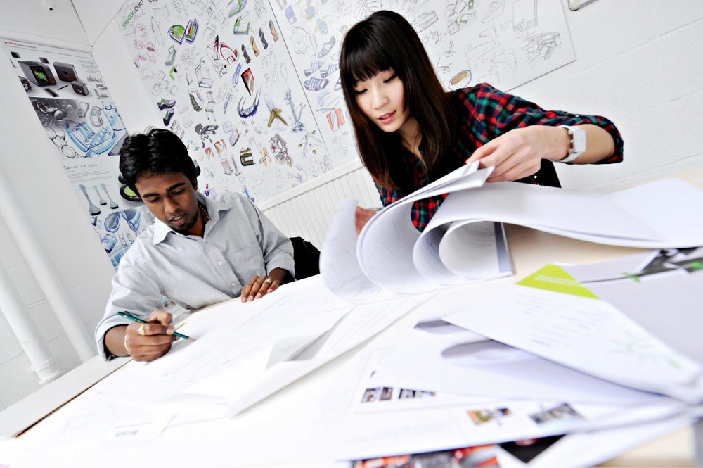 Image of two people looking at their design work. One is sketching, and the other is going through a work book of designs. 