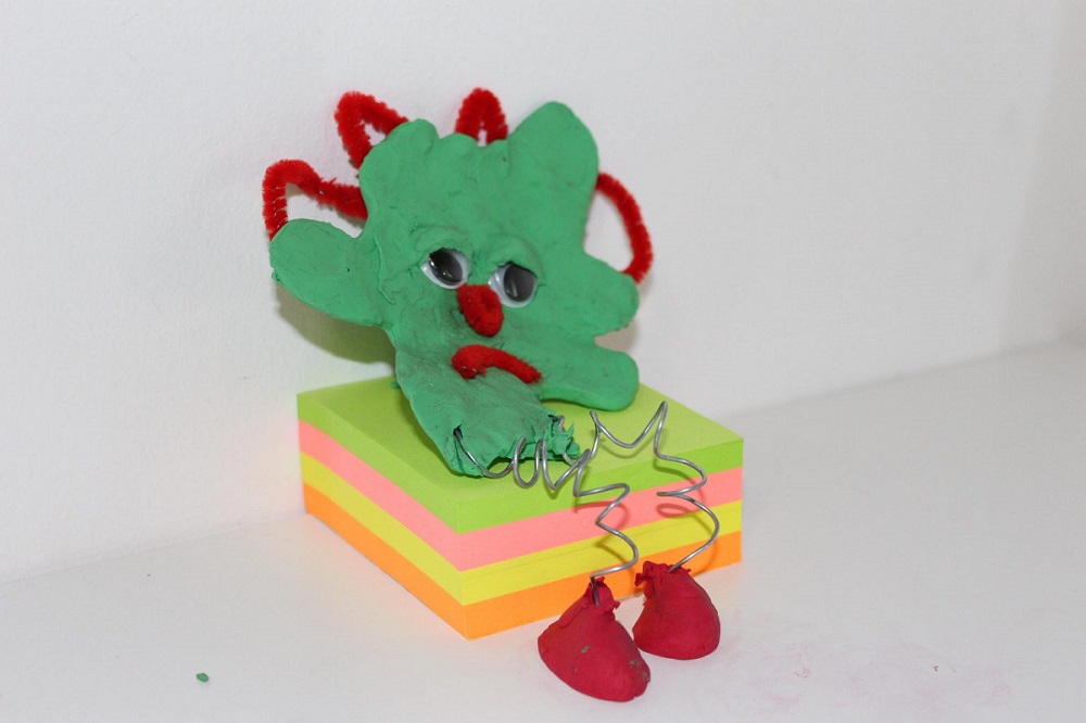 A green clay model Emma has created with plastic eyes and fuzzy red pipe cleaners for facial definition. It has metal spring legs and red clay feet. It is leaning on a stack of multi-coloured sticky notes.  