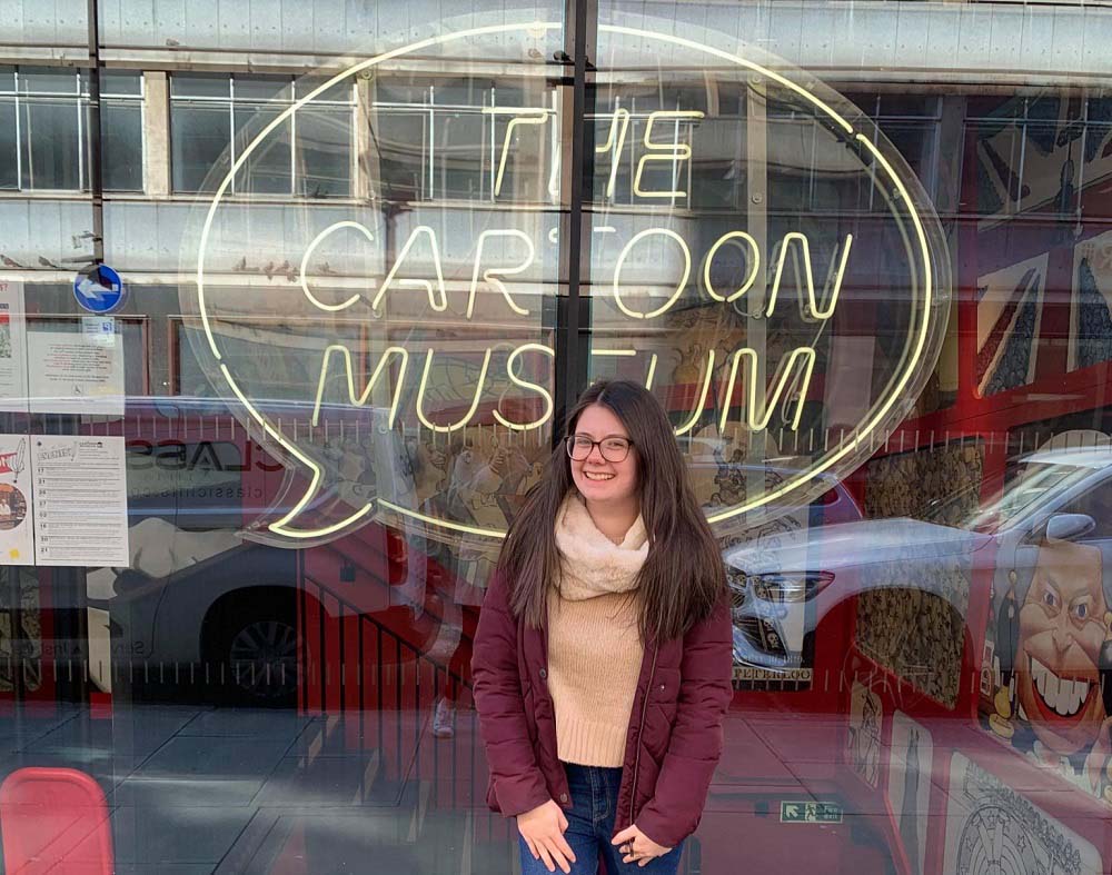 Emma standing in front of an yellow sign of a speech bubble with the words 'the cartoon museum' in it. 