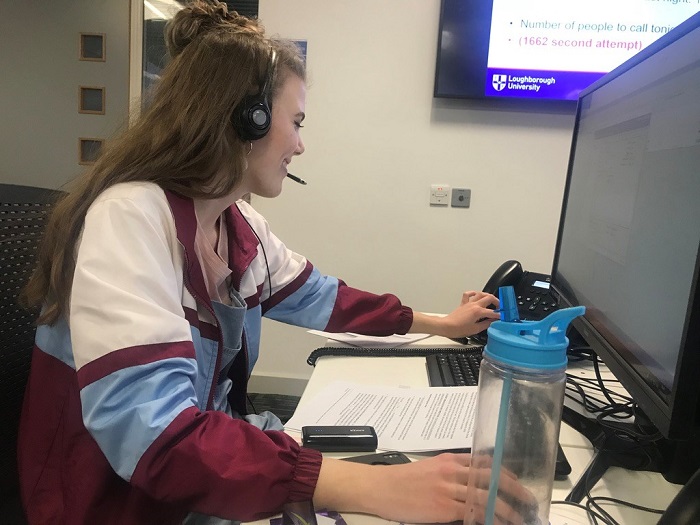 Caroline working on a computer with a headset on. In the photo she is going to answer the phone which is to the left of the computer which is in front of her. 