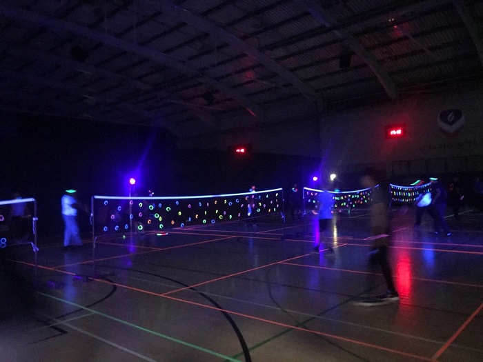 Students participating in UV Badminton in a sports hall. The students have brightly coloured clothes on and the nets have glow sticks and vibrant tape on them, which reflects off the UV light. The sports hall is dark as the main lights are turned off to give maximum effect.  