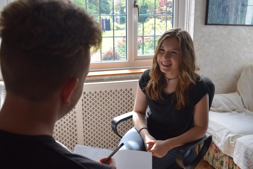 Lauren sitting on a chair chatting to her brother who is writing notes in a notepad.  