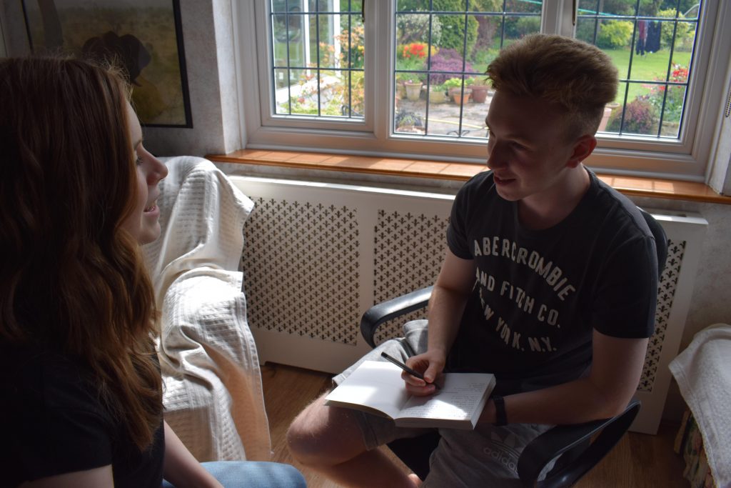 Lauren and her brother sitting opposite each other in chairs. Her brother is asking her questions and writing notes in a notepad. 