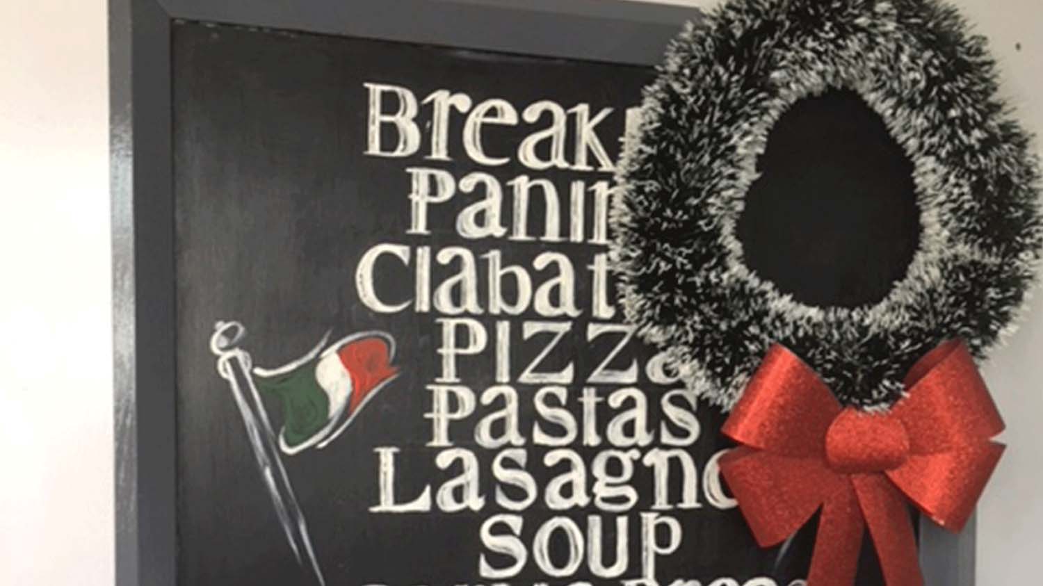 A sign outside the Italian in Loughborough which lists food items that they serve handwritten in a list. They are breakfast, panini, ciabatta, pizza, pastas, lasagna, and soup. The sign also has an Italian flag drawn on it and a green and red wreath hung on it to the right side.   