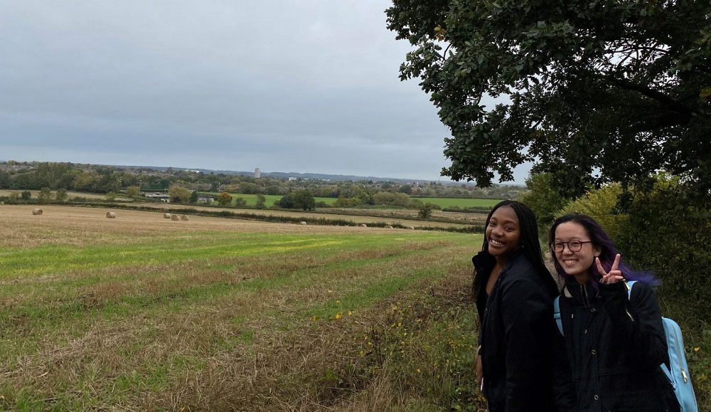 Nyasha and a friend smiling towards the camera in a field that overlooks the countryside. 