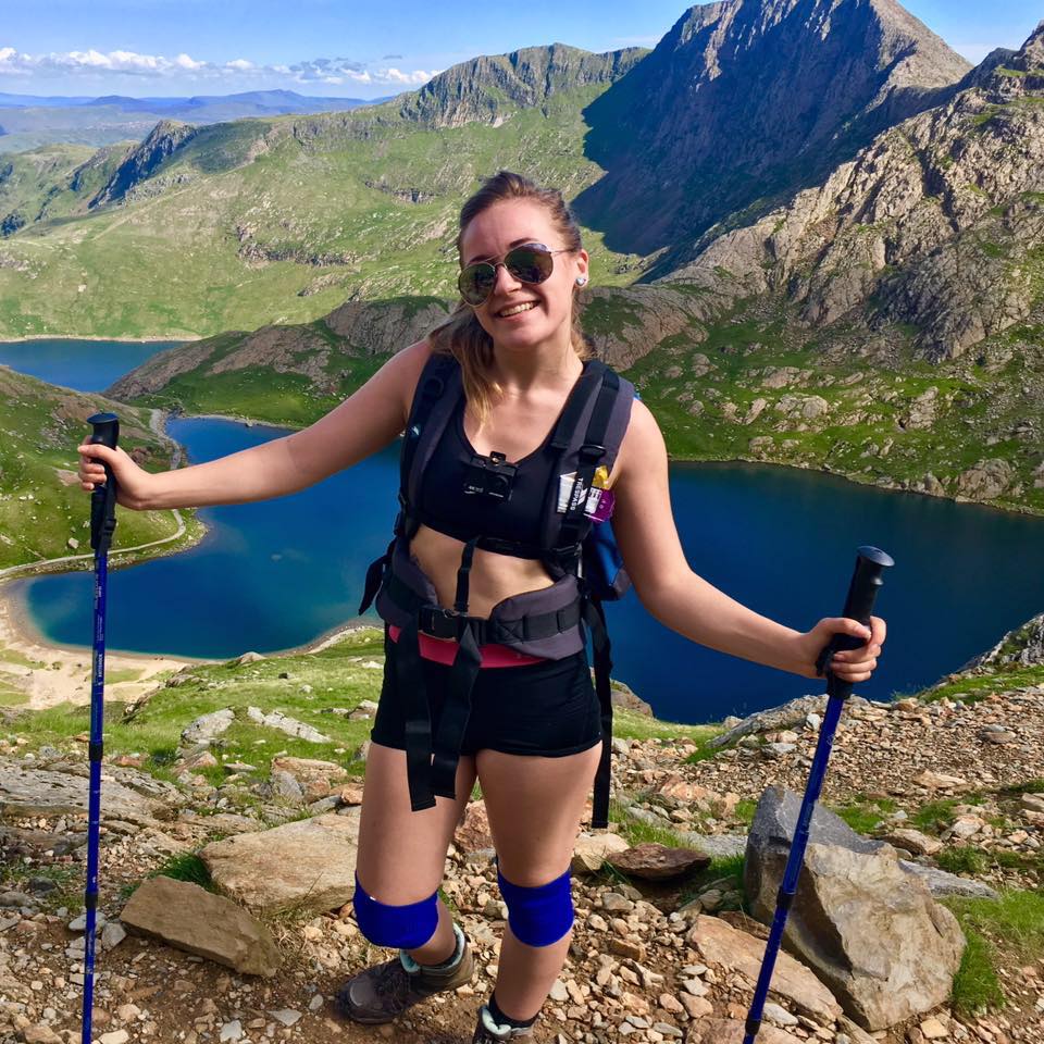 Jess hiking on an expedition. She is standing at the top of a mountain with a view of another mountain and a lake in the background. 