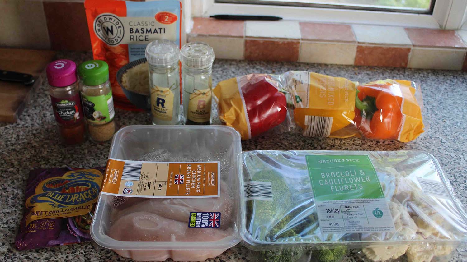 Photo of some ingredients to make a meal. There is chicken, peppers, rice, broccoli and cauliflower alongside herbs and spices.  