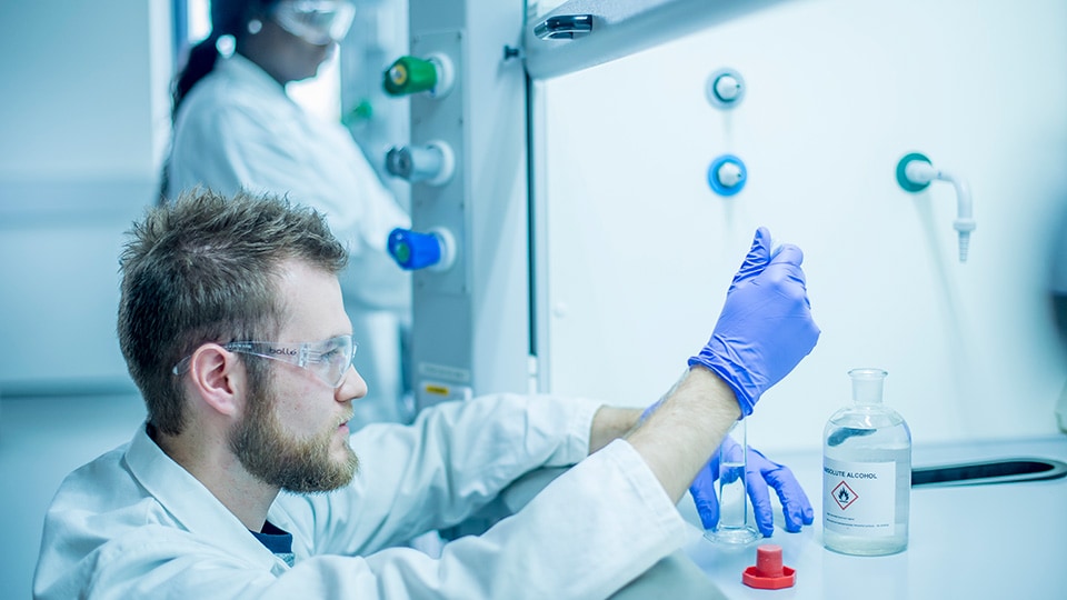 Two people are working in a lab and they are both wearing lab coats, protective eyewear and gloves. One person is filling a test tube with Absolute Alcohol using a pipette.
