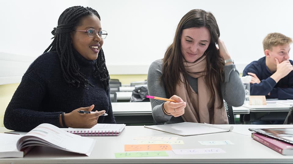 Two students sat at a table with books, notepaper, pens and post it notes.