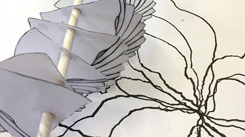 Paper sculpture prototype with layered cut-out shapes mounted on a rod beside a hand-drawn design sketch.