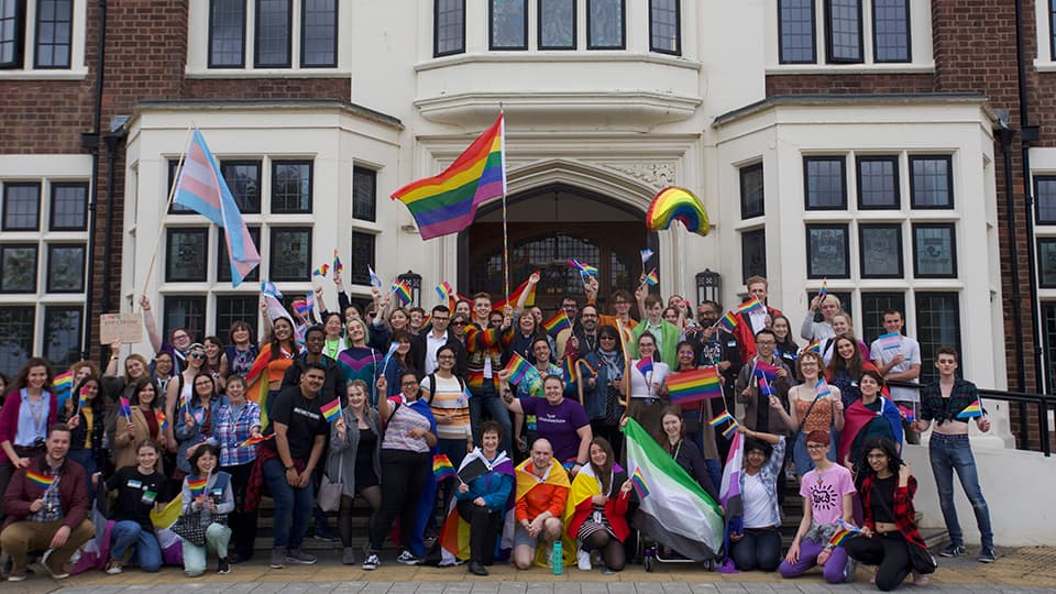 a group of people holding Pride flags and banners on the steps outside the Hazlerigg building