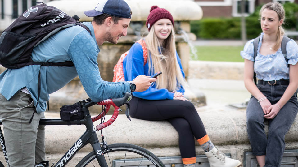 One student on a bike and two students sat by the fountain on campus.