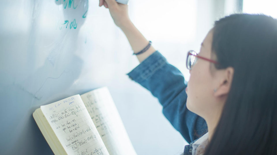 A student, holding a pen and a notepad, and writing on a whiteboard