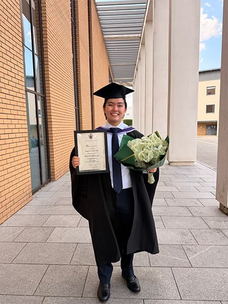 Laurel wearing a graduation gown and cap, holding a bouquet of flowers and a certificate.